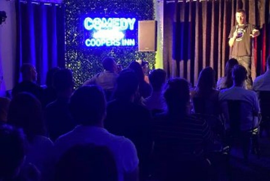 Stand-up comedian holding a microphone on a stage in front of curtains, a big screen and seated audience.